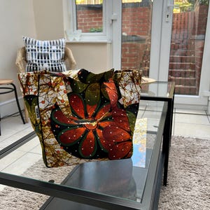 Puede incluir: Un bolso tote colorido con un estampado floral en tonos naranja, verde y negro. El bolso es de tela y está sobre una mesa de cristal. El diseño del bolso presenta una gran flor estilizada.