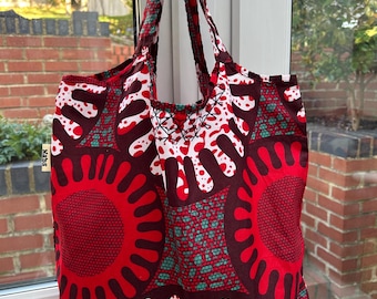 Pode incluir: Uma vibrante sacola com um padrão arrojado e repetitivo em vermelho, branco e verde-azulado. O design apresenta grandes explosões solares estilizadas e formas orgânicas sobre um fundo vermelho escuro. A sacola tem alças estampadas.