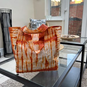 May include: A tie-dye patterned tote bag in shades of orange and white sits on a glass-topped coffee table. The bag has two handles and the word "Mahs" is visible on the side. The bag is open, revealing its interior.