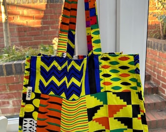 Pode incluir: Uma bolsa tote colorida com um design de patchwork que apresenta padrões geométricos em amarelo, laranja, azul, verde e preto. A bolsa tem uma etiqueta preta com a palavra "NABS" impressa nela.