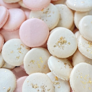 May include: A close-up of a pile of pink and white macarons with gold leaf accents. The macarons are arranged in a random pattern, with some of the pink macarons on top of the white macarons.