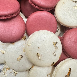 May include: A close-up of a pile of macarons. Some are white with gold flakes and some are a deep pink color.
