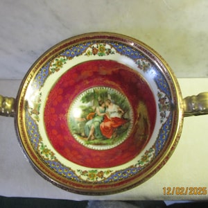 May include: A gold-rimmed porcelain bowl with a red and white floral design. The bowl has two gold handles and a central image of two women in a garden setting.