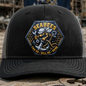 May include: A black baseball cap featuring a hexagonal patch on the front. The patch displays a bee wearing a hard hat, holding a wrench and an anchor, with the text SEABEES and CAN DO WILL DO DONE in yellow lettering against a dark background.