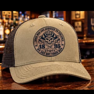 May include: A tan trucker hat with a dark brown mesh back and a circular graphic on the front. The graphic features a skull wearing a pirate hat, crossed swords, the text USN, the numbers 18 and 93, and the phrase SOME MEN ARE OPPOSED TO VIOLENCE WE ARE NOT THOSE MEN.