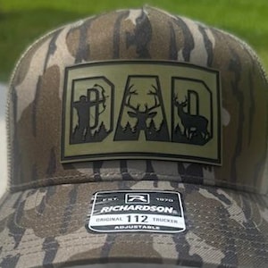 May include: A camouflage trucker hat with a mesh back and a curved brim. The front patch reads "DAD" with a deer and hunter silhouette. The hat is adjustable and features the Richardson brand logo.