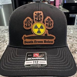 May include: A black and brown trucker hat with a leather patch that says "Death From Below". The patch features a submarine with three anchors and a yellow radiation symbol. The hat is adjustable and has a Richardson logo.