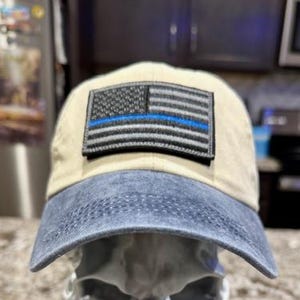 May include: A beige and blue baseball cap featuring a patch with a black and gray American flag design and a blue stripe. The cap is displayed on a silver skull-shaped decorative object.