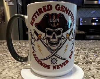 May include: White ceramic coffee mug with a black handle. The mug has a graphic of a skull wearing a pirate hat with the text "Retired Genuine Legends Never Die" and "USN" on the hat.