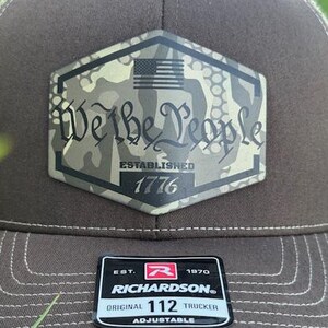 May include: Brown trucker hat with a camouflage patch featuring the text "We the People" and an American flag. The hat includes the text "Established 1776" and a Richardson patch.