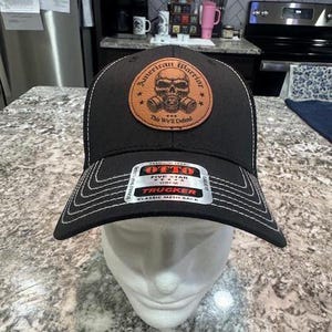 May include: Black trucker hat with a brown leather patch that says "American Warrior" with a skull and crossbones design. The patch also says "This We'll Defend". The hat has a white mesh back and a label that says "Otto Five Star OS/M Trucker Classic Mesh Back".