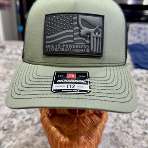 May include: Olive green trucker hat with a black mesh back. Features a black patch with a skull, American flag, and the text "EVIL IS POWERLESS IF THE GOOD ARE UNAFRAID." The hat is adjustable and has a Richardson tag.
