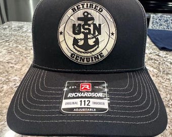 May include: A black trucker hat with a gray mesh back. The front of the hat features a black and white anchor with the text "RETIRED USN GENUINE". The hat also has a black and white label that reads "EST. 1870 RICHARDSON ORIGINAL 112 TRUCKER ADJUSTABLE".