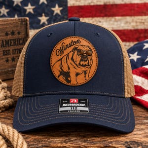 May include: A navy blue trucker hat with a tan mesh back and a circular tan leather patch featuring an illustration of a bulldog wearing American flag sunglasses with the text Winston above it. A black sticker on the brim reads Richardson 112 Trucker.