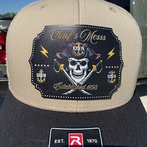 May include: A tan and black baseball cap with a patch featuring a skull wearing a pirate hat, crossed swords, and the text "Chief's Messs". The patch also includes the text "Established 1893" and "USN". The cap is made by Richardson.