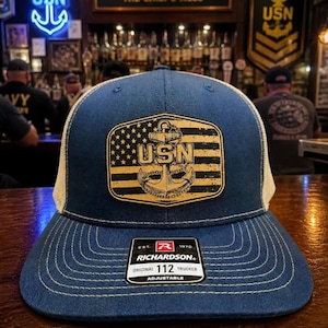 May include: A navy blue trucker hat with a tan mesh back. The front features a patch with the US flag, the letters "USN", and an anchor. The hat is adjustable and has the Richardson 112 logo.