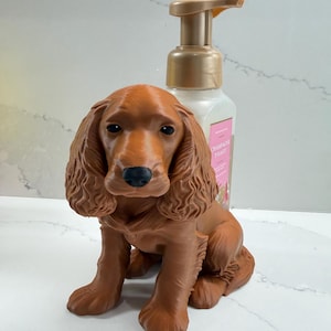 May include: A brown spaniel dog figurine with long ears and a black nose positioned in front of a square soap dispenser bottle with a gold pump. The bottle label features pink text that reads Champagne Toast in a clear, readable font.