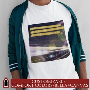 May include: White t-shirt with a graphic of a blurred city lights scene and a quote from Deuteronomy 33:12: "The beloved of Yahweh will dwell in safety, secure in His, God shields him all day long. He rests between the shoulders of the Lord." The shirt is customizable with comfort colors and Bella + Canvas.