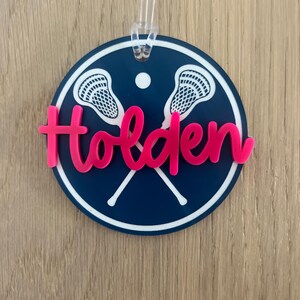 May include: A blue and white circular lacrosse themed ornament with the name Holden in pink lettering.