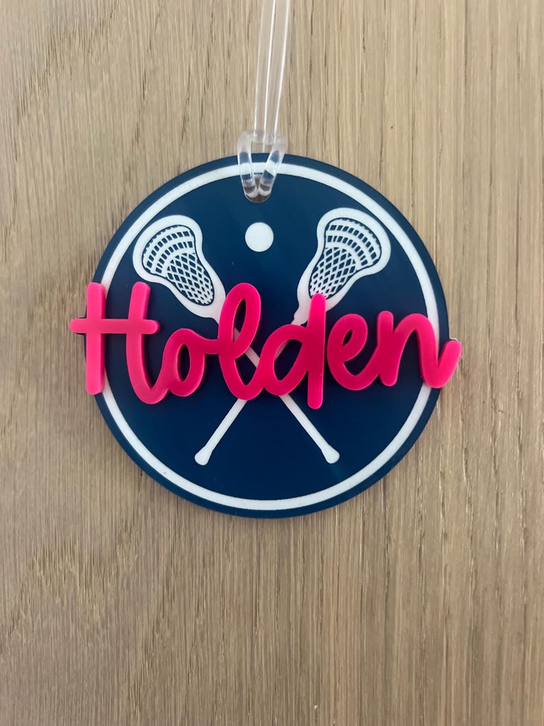May include: A blue and white circular lacrosse themed ornament with the name Holden in pink lettering.