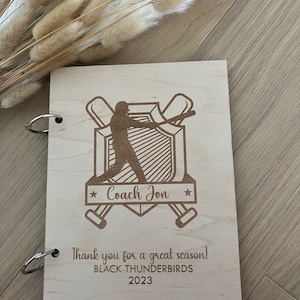 May include: A wooden notebook with a baseball theme. It features a baseball player silhouette, bats, and a shield with the text "Coach Jon". The notebook also has the text "Thank you for a great season! BLACK THUNDERBIRDS 2023".