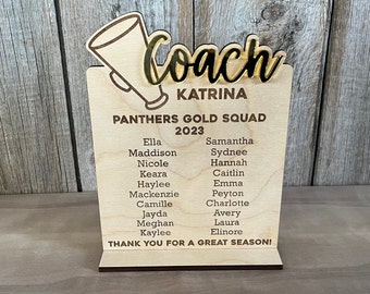 May include: A light-colored wooden plaque featuring a laser-engraved megaphone and the word Coach in black script. It lists Panthers Gold Squad 2023, the name Katrina, a list of names in two columns, and the text THANK YOU FOR A GREAT SEASON!