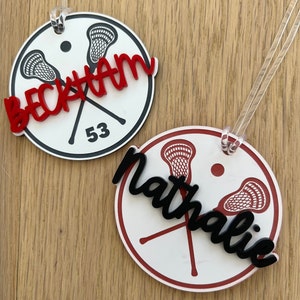 May include: Two white round lacrosse themed ornaments with black and red lacrosse sticks and a black and red name. The first ornament says "Beckham 53" in red. The second ornament says "Nathalie" in black.
