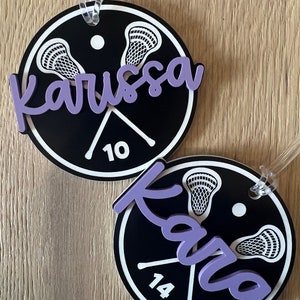 May include: Two black and white round lacrosse themed luggage tags with purple lettering. The top tag says "Karissa 10" and the bottom tag says "Karisa 14".