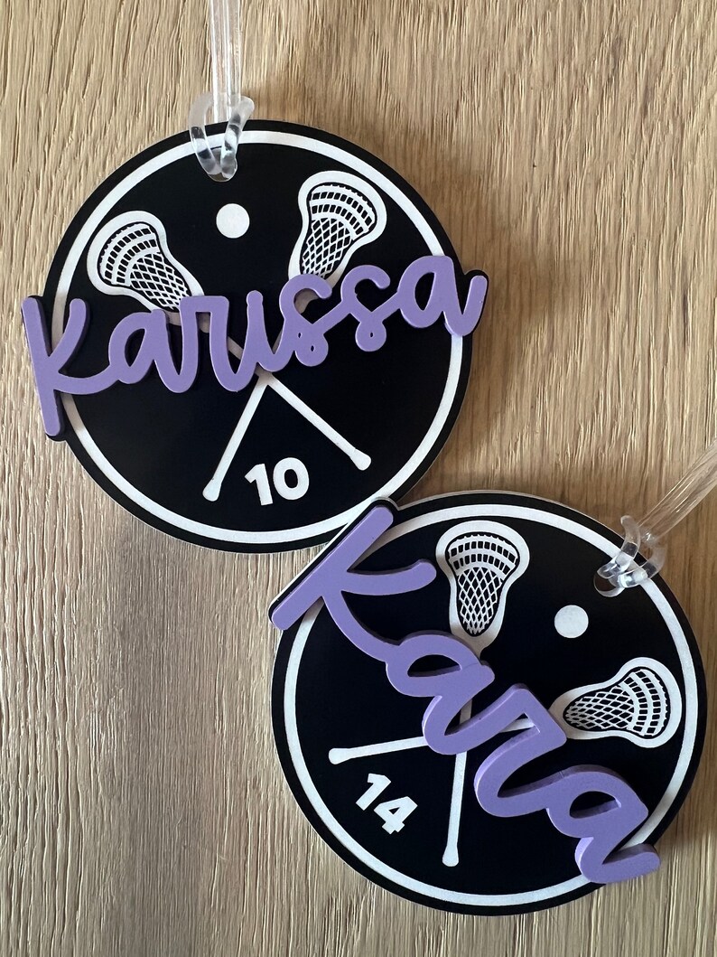 May include: Two black and white round lacrosse themed luggage tags with purple lettering. The top tag says "Karissa 10" and the bottom tag says "Karisa 14".