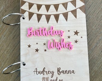May include: A wooden birthday wishes scrapbook with a light wood finish. The cover features a banner design, pink "Birthday Wishes" text, and star accents. The name "Audrey Banna" and the year "2010 and on..." are also included. The scrapbook is held together with silver rings.