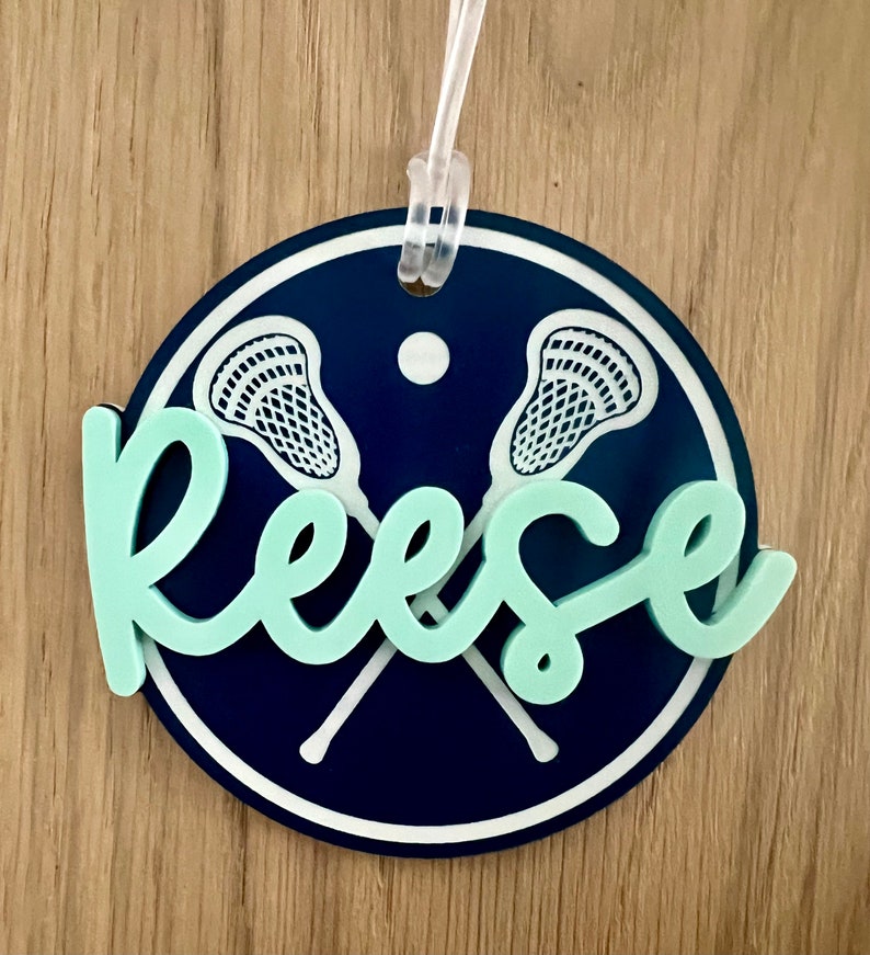 May include: A round, blue and white lacrosse themed luggage tag with the name "Reese" in light green.