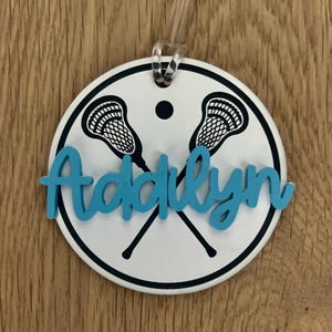 May include: A white round luggage tag with a black outline and two lacrosse sticks crossed in the center. The name "Addilyn" is written in blue cursive letters.
