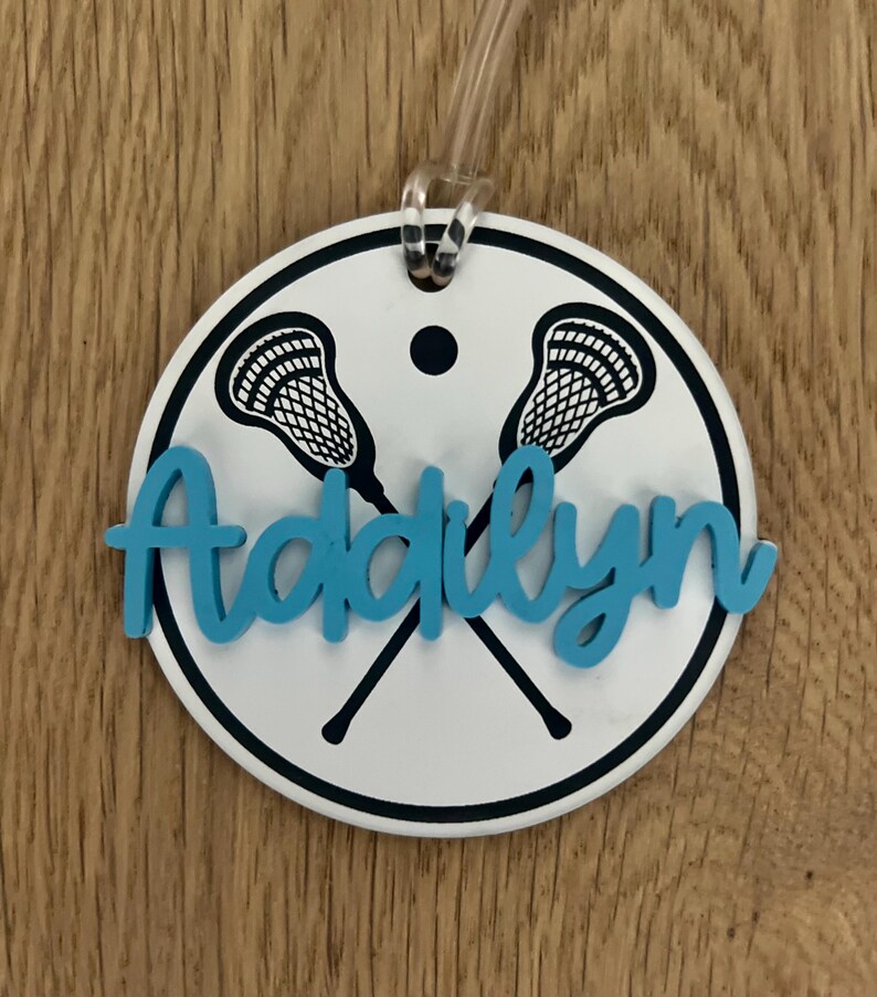 May include: A white round luggage tag with a black outline and two lacrosse sticks crossed in the center. The name "Addilyn" is written in blue cursive letters.