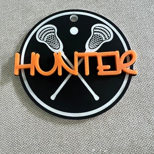 May include: Black and white lacrosse stick design on a round black background with a white border. The name "HUNTER" is written in orange across the sticks.
