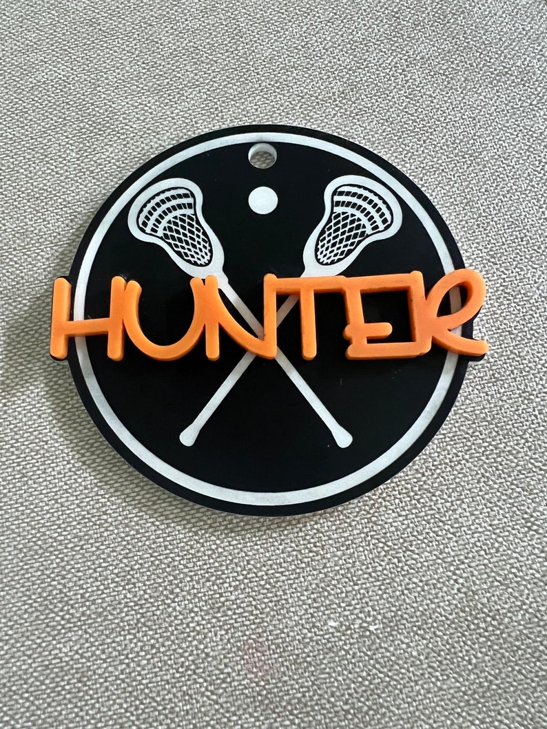 May include: Black and white lacrosse stick design on a round black background with a white border. The name "HUNTER" is written in orange across the sticks.