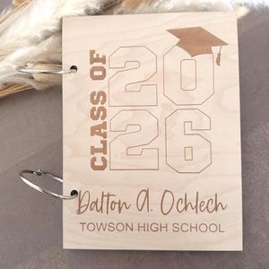 May include: A light brown wooden graduation album with silver binder rings. The cover is engraved with "CLASS OF 2026", a graduation cap, the name "Dalton A. Ochlech", and "TOWSON HIGH SCHOOL".