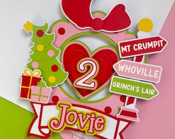 May include: A festive cake topper with a red bow, a green Christmas tree, and a banner that says "Jovie". The topper features a heart with the number "2" and directional signs pointing to Mt. Crumpit, Whoville, and Grinch's Lair. The color scheme includes red, green, pink, and yellow.