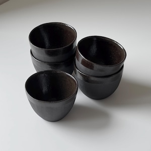 May include: Six small, black ceramic bowls stacked on a white surface. The bowls have a smooth, glossy exterior and a dark interior. The bowls are arranged in a slightly offset stack. These bowls are likely for serving small portions of food.