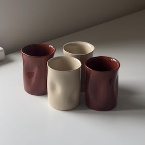 Könnte beinhalten: Ein Set aus vier Keramikbechern mit glatter, glänzender Oberfläche. Zwei Becher sind in einem tiefen Rotbraun gehalten, zwei in einem hellen Cremeton. Jeder Becher weist eine einzigartige, leicht eingedellte, organische Form auf.