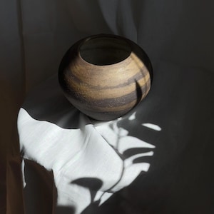 Peut inclure: Un vase sphérique en céramique fait à la main, avec une large ouverture. Il présente des bandes horizontales de tons bruns et beiges. Le vase est posé sur un fond blanc, avec des ombres projetées par la lumière du soleil.