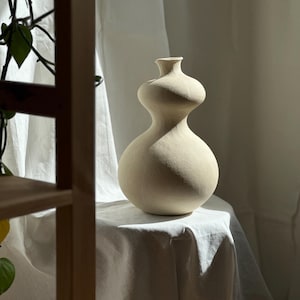 Op de afbeelding: Een matte, gebroken witte keramische vaas met een opvallend dubbel-kalebas silhouet, voorzien van een brede ronde basis die taps toeloopt naar een smalle hals en een kleine uitlopende rand, met een gladde, ongeglazuurde textuur.
