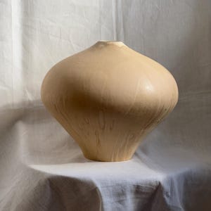 Könnte beinhalten: Eine breite, bauchige Keramikvase mit einer glatten, matten beigen Oberfläche. Das Gefäß hat eine schmale, kreisförmige Öffnung an der Oberseite und einen verjüngten Boden, mit dezenten, vertikalen, helleren tropfenartigen Texturen.