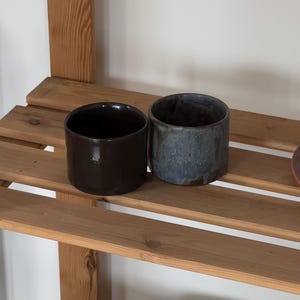 Könnte beinhalten: Zwei zylindrische Keramikbecher stehen nebeneinander. Der linke Becher hat eine glatte, glänzende dunkelbraune Oberfläche. Der rechte Becher weist eine strukturierte, matte Oberfläche mit gesprenkelten Blau-, Grau- und Dunkelbrauntönen auf.