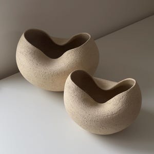 Op de afbeelding: Twee beige keramische kommen met een getextureerde, zanderige buitenkant en een onregelmatige, organische randvorm. De kommen hebben een afgeronde, asymmetrische vorm met een glad binnenoppervlak en een neutrale kleur en tactiele afwerking.