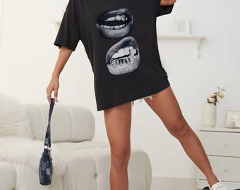 May include: Black oversized t-shirt featuring a graphic of two sets of lips with braces. The top is worn by a person. The person is wearing white sneakers and holding a small denim purse. The background includes a white couch and a white rug.