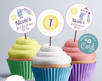 May include: Three cupcakes with colorful frosting and cupcake liners. Each cupcake has a round, white paper topper with a sun and a purple astronaut. The toppers say "Nicole's First Trip Around the Sun" and "1".