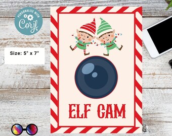 May include: Printable Elf Cam sign with two elves holding Christmas lights, a camera lens, and the text "ELF CAM" in red letters. The sign has a red and white candy cane striped border.