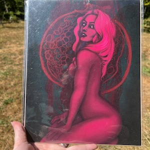 May include: A digital illustration of a woman with pink hair and skin, sitting with her legs crossed. She is wearing a dark background with a circular design behind her.
