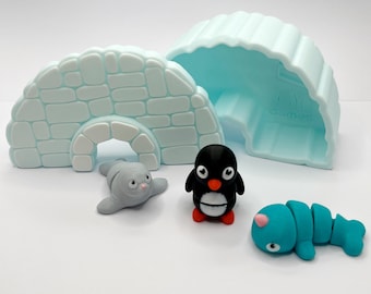 Puede incluir: Un juego de juguetes con un iglú azul claro y una cueva de hielo. El juego incluye un pingüino blanco y negro con pico y patas rojos, una foca gris y un pez azul segmentado. El iglú está hecho de bloques individuales.