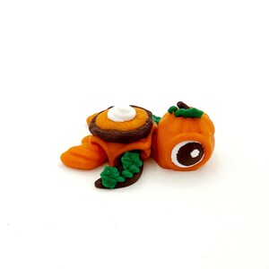 以下が含まれることがあります： 緑の葉と茶色の甲羅を持つ、小さなオレンジ色のカボチャの亀の置物。亀の甲羅の上には、ホイップクリームのような白い渦巻きがある。